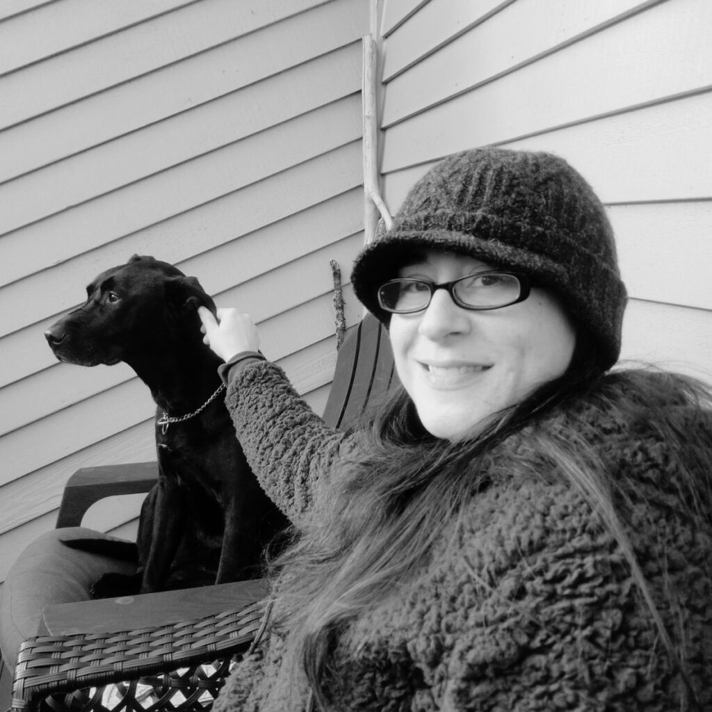 Alyda, wearing a black hat and cozy jacket, smiles while petting a black dog sitting beside her on a chair, with a wooden wall in the background.